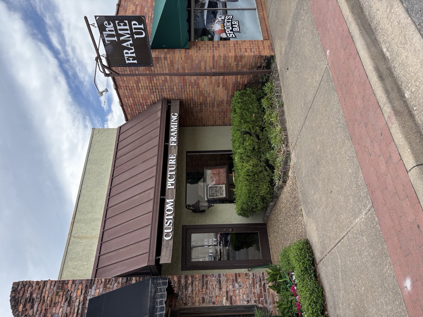 The Frame Up storefront on Vienna Street in downtown Ruston, Louisiana — brown standing-seam awning, ‘Custom Picture Framing’ lettering, hanging Frame Up sign on red brick